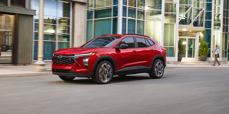 Red 2026 Chevy Trax parked in front of a modern building, highlighting its compact design, bold styling, and urban versatility.